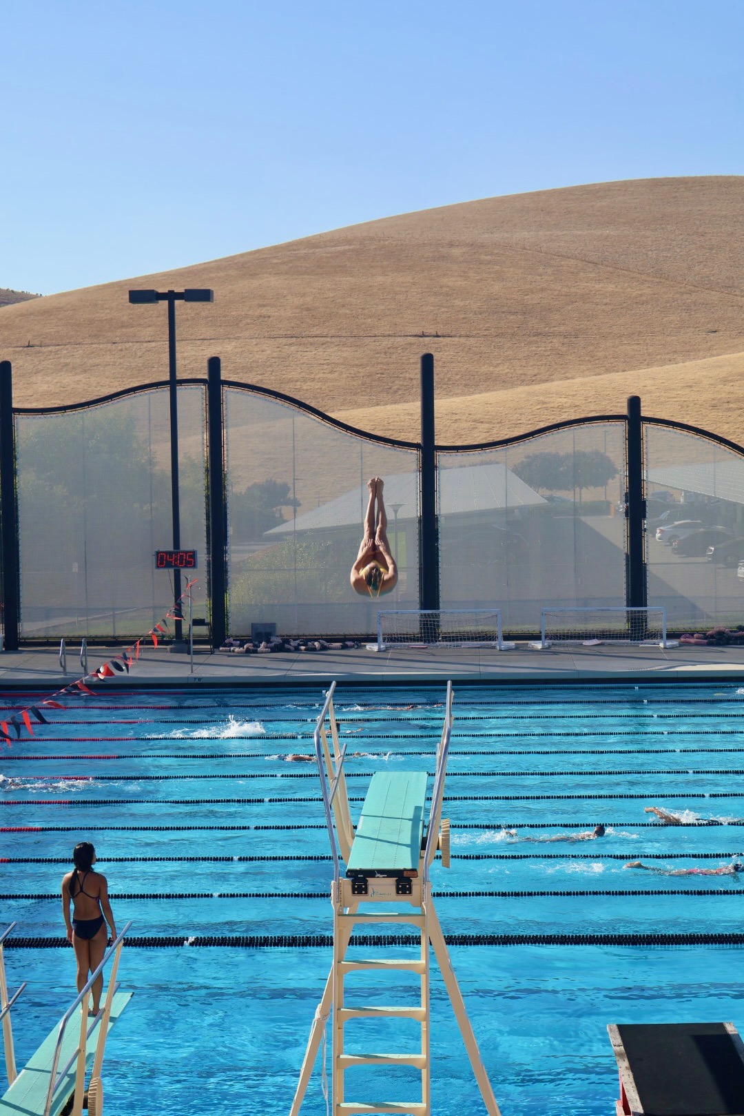 Junior Olympic diver in a vertical pike entry high above the pool with the Livermore hills behind